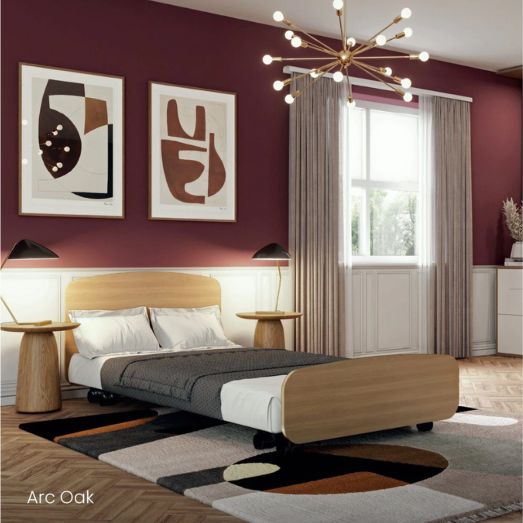 Modern bedroom featuring an Accora Contesa® FloorBed® Hi/Low Full Electric Bed for stylish fall prevention, plus a geometric rug, abstract art on a maroon wall, wooden side tables with black lamps, beige curtains, and a gold sputnik chandelier.