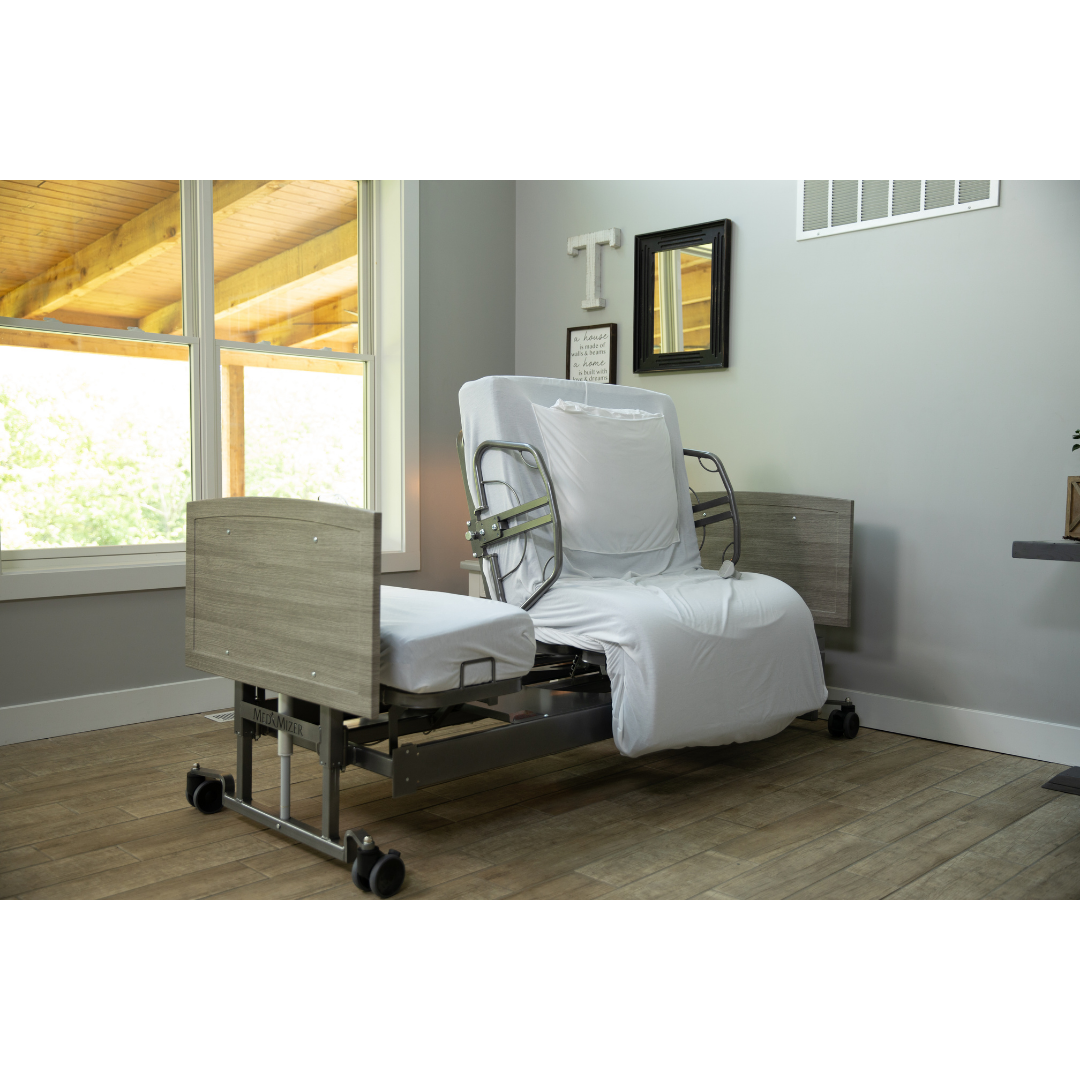 The Med-Mizer ActiveCare Rotating Hospital Bed, featuring light wood panels and white bedding, is placed in a bright, tidy room with large windows and décor—an ideal choice for comfortable home care.