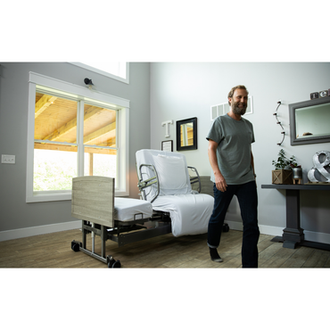A man with a beard walks across a bright, modern room with large windows; behind him is the Med-Mizer ActiveCare Rotating Hospital Bed featuring white bedding and wooden accents.