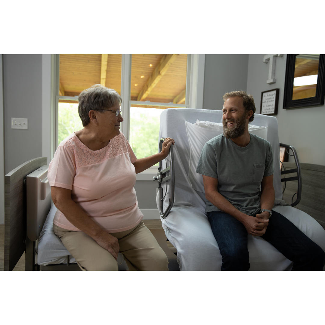 An older woman and a bearded man smile and chat while sitting on Med-Mizer ActiveCare Rotating Hospital Beds in a bright room with natural light streaming through a large window behind them.