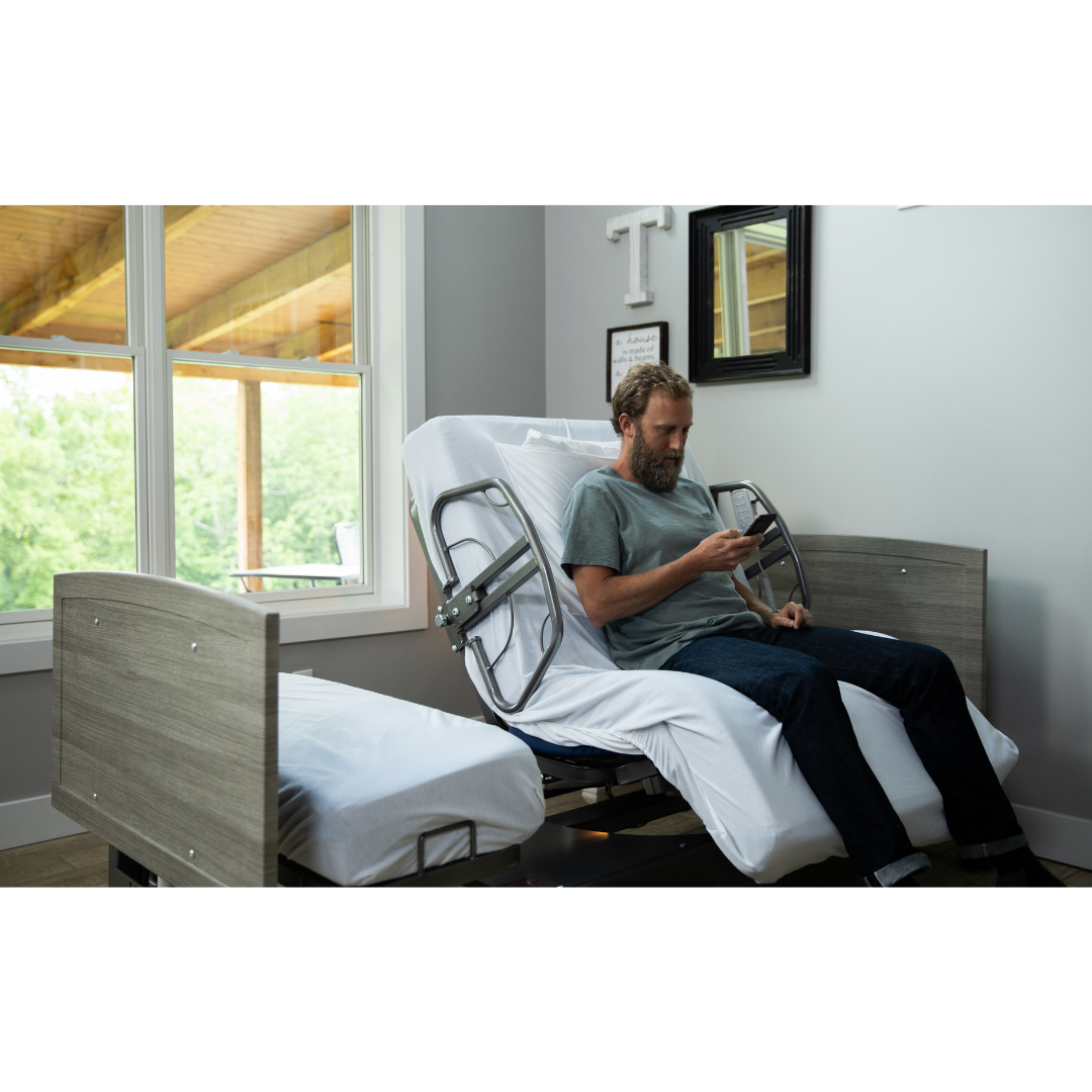 A man sits on a Med-Mizer ActiveCare Rotating Hospital Bed in a bright room, partially elevated by large windows with greenery outside, while he looks at his phone.