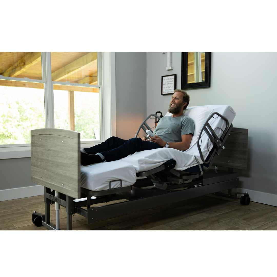 A man reclines in a bright, spacious room on the Med-Mizer ActiveCare Rotating Hospital Bed, holding a remote control and appearing relaxed.