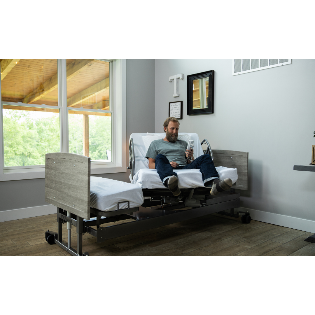 A man relaxes on a Med-Mizer ActiveCare Rotating Hospital Bed in a bright room with large windows, wooden floors, and wall decor as he looks at his phone.