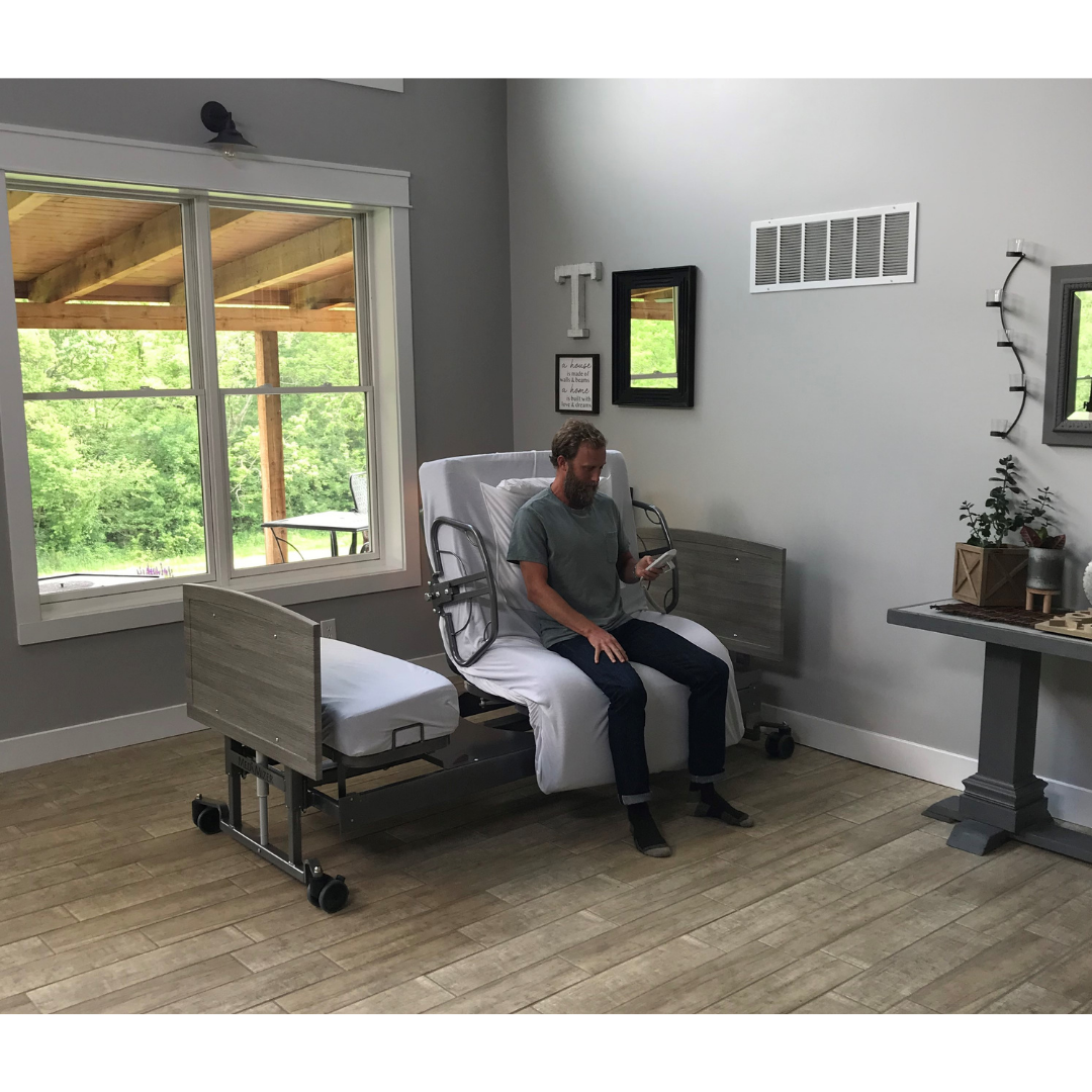 A man sits on the edge of a Med-Mizer ActiveCare Rotating Hospital Bed in a bright room with wood floors, large windows, and home decor, looking down at something in his hand.