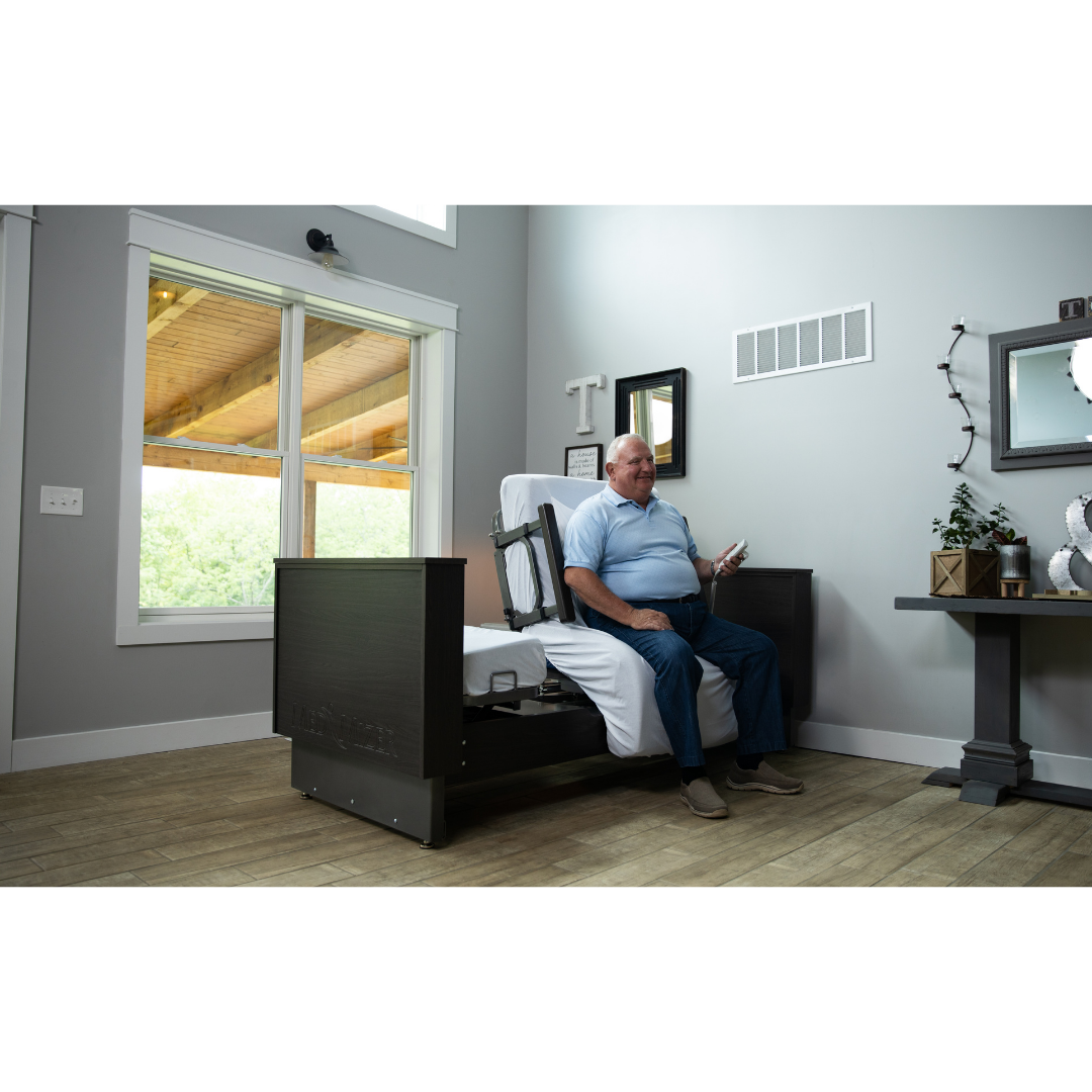 An older man sits on the edge of a Med-Mizer ActiveCare Rotating Hospital Bed in a bright, modern bedroom with large windows, wooden floors, and decor on a side table, holding a remote control.