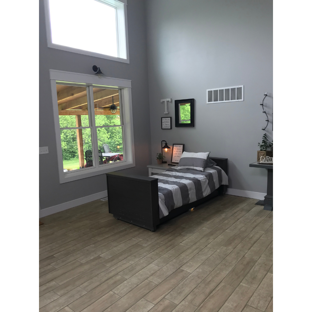 A modern minimalist bedroom with gray walls and light wood floors features a Med-Mizer ActiveCare Rotating Hospital Bed dressed in gray and white striped bedding. Large windows bring in sunlight and a small nightstand stands beside the bed.