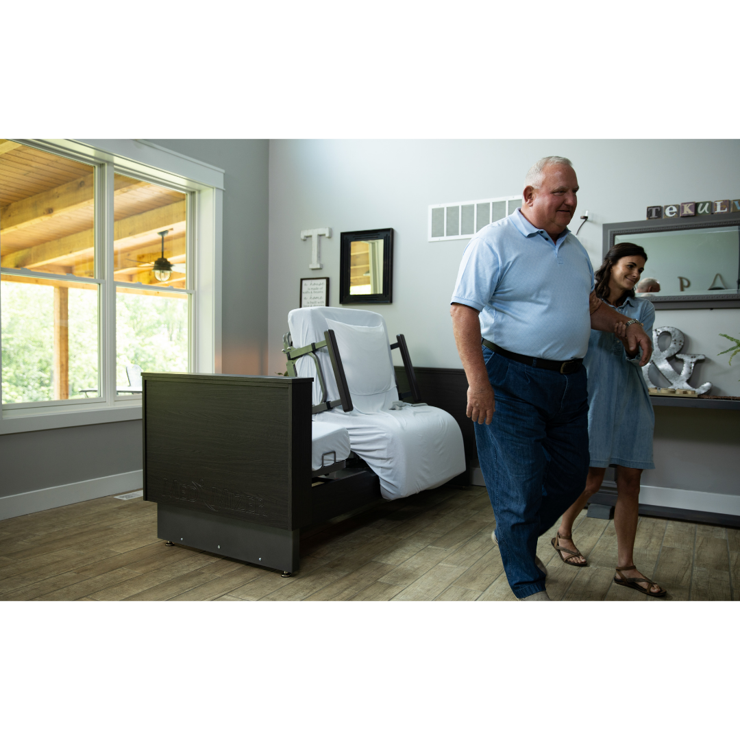 An older man and a woman walk together in a bright, modern bedroom with large windows, featuring the Med-Mizer ActiveCare Rotating Hospital Bed.