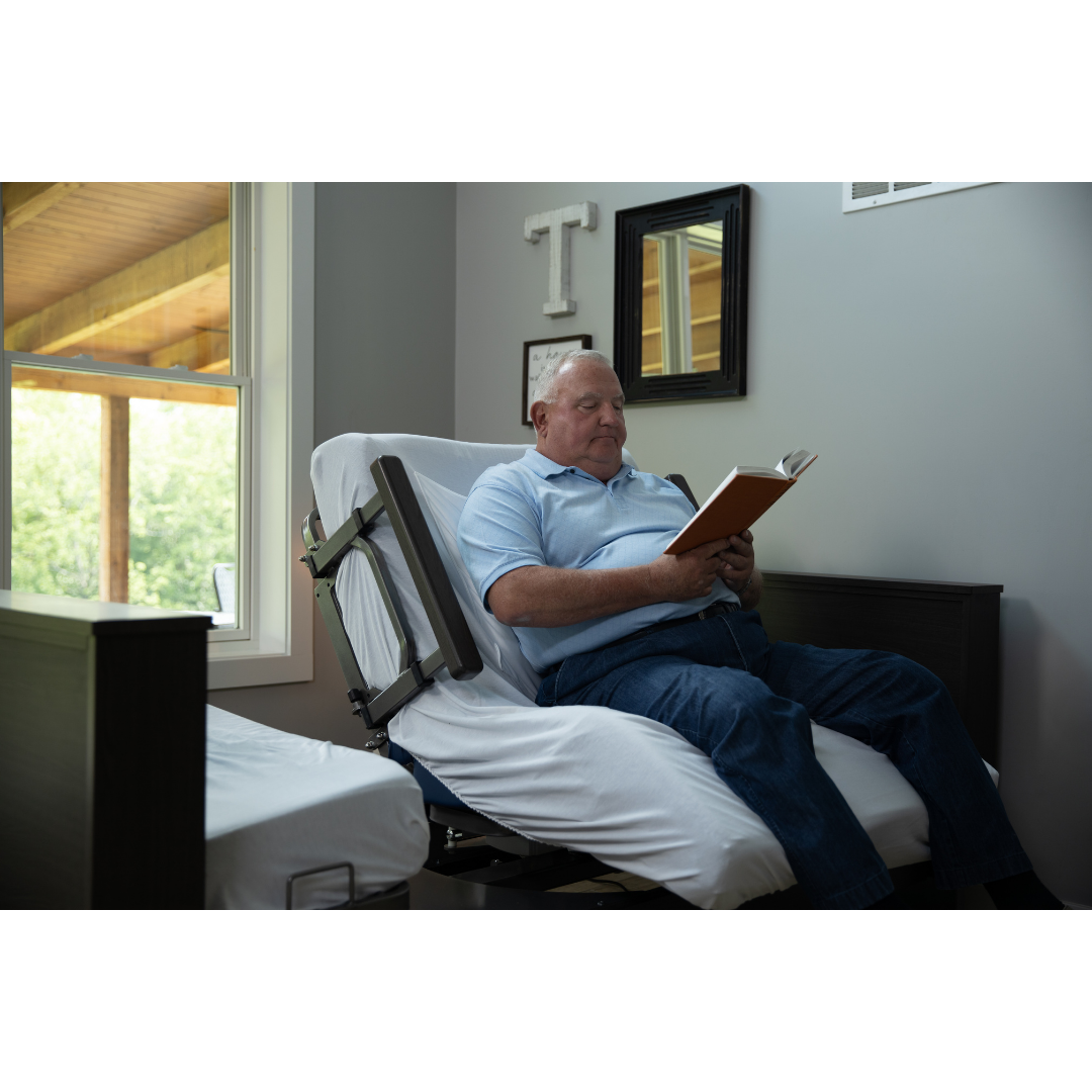 An older man sits upright in a Med-Mizer ActiveCare Rotating Hospital Bed, reading a book in a bright room with a window, black mirror, and wall art.