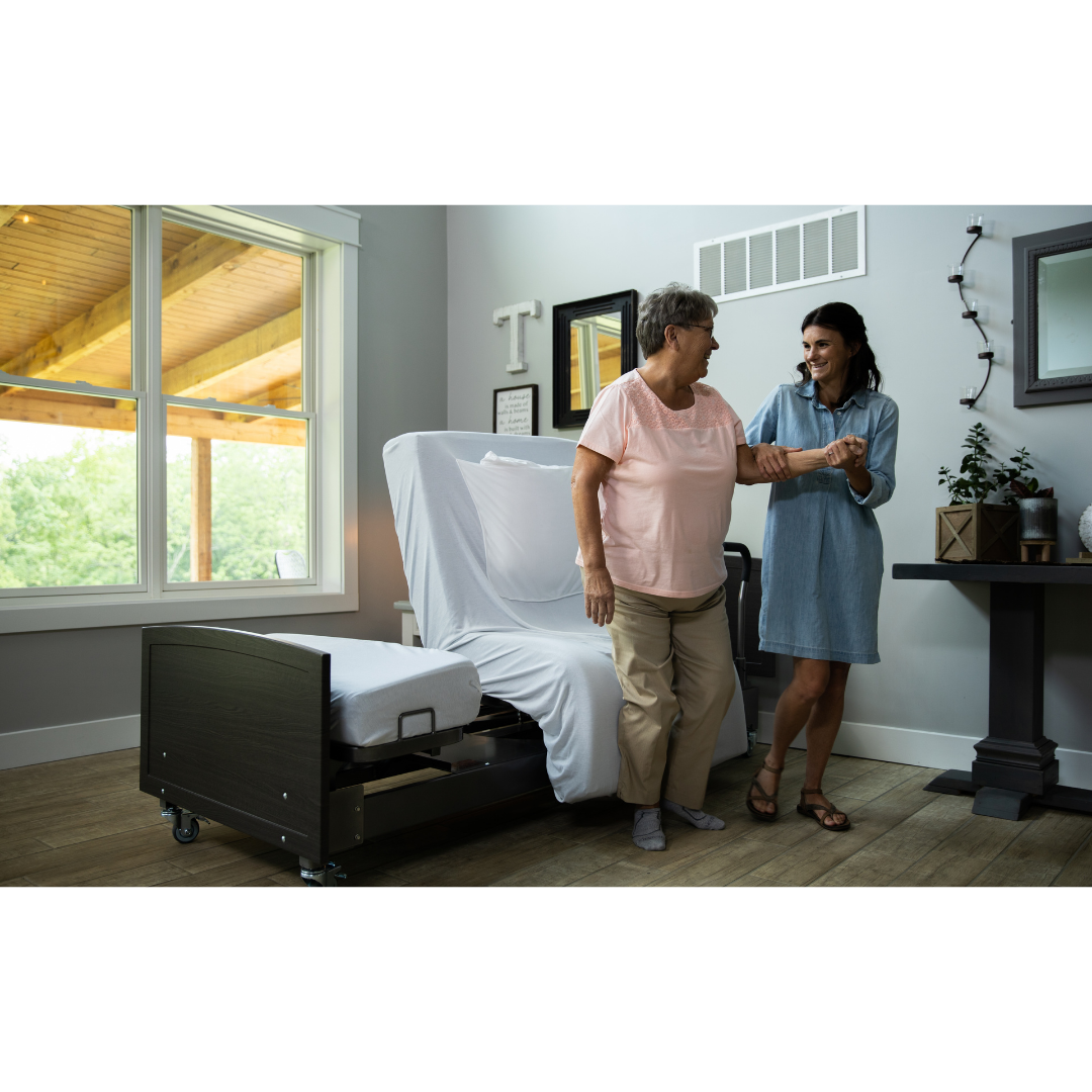 An older woman stands beside a Med-Mizer ActiveCare Rotating Hospital Bed, smiling and holding hands with a younger woman in a sunlit room with wooden floors and large windows.