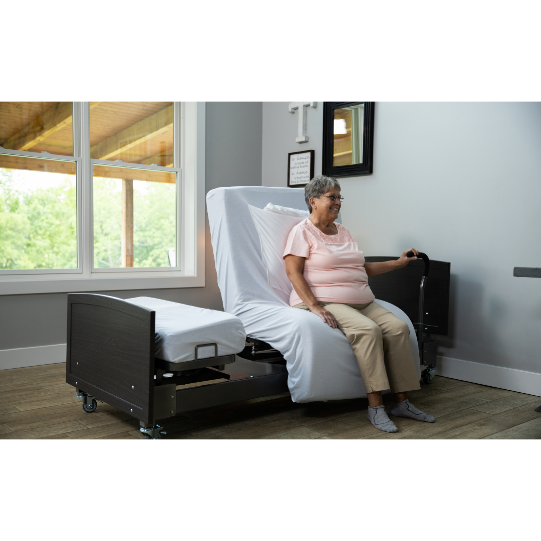 An older woman sits on the edge of a Med-Mizer ActiveCare Rotating Hospital Bed in a bright room with large windows, holding a support rail and smiling.