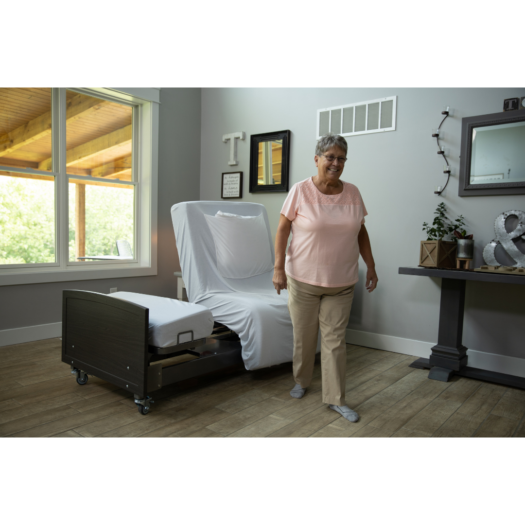 An older woman in glasses, a pink blouse, and beige pants smiles beside a Med-Mizer ActiveCare Rotating Hospital Bed in a bright bedroom with wooden floors and large windows.