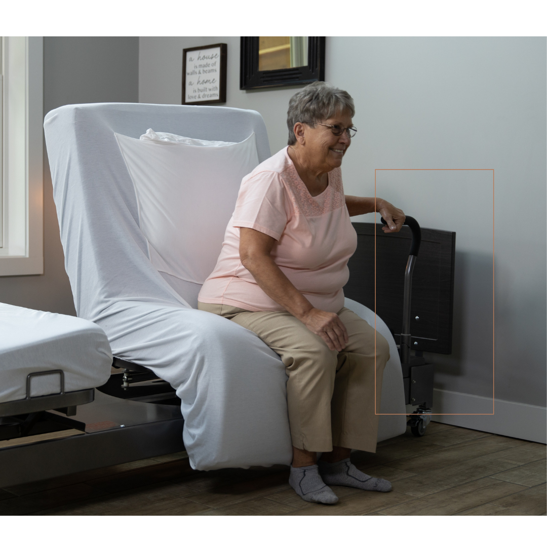 An older woman sits on the edge of a Med-Mizer ActiveCare Rotating Hospital Bed, smiling with one hand on a black walker. She wears glasses, a pink top, and beige pants in a softly lit room with wooden flooring.