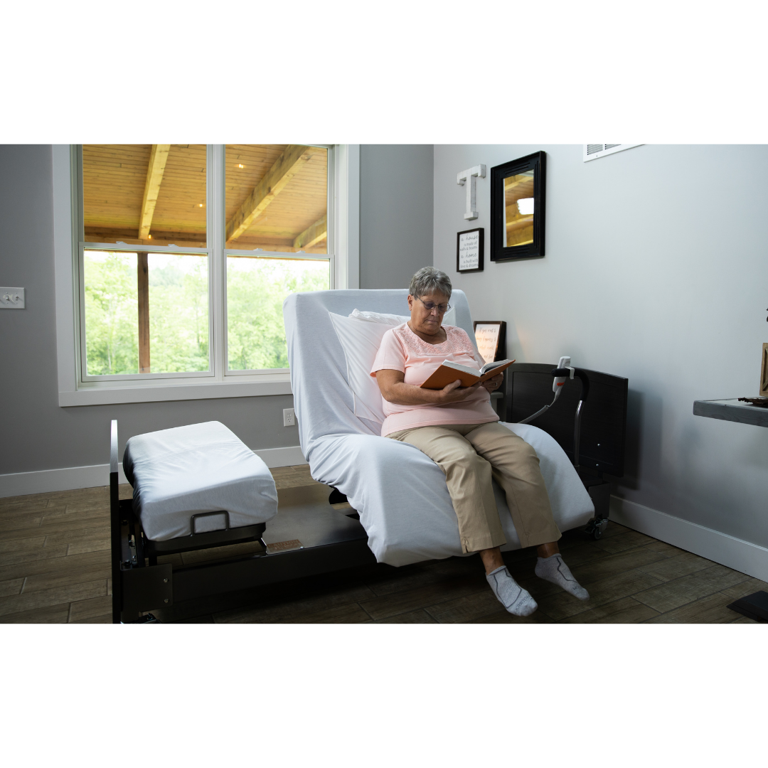 An older woman reads a book on the Med-Mizer ActiveCare Rotating Hospital Bed in a bright room, wearing glasses, a pink shirt, and beige pants as sunlight shines through the large window behind her.