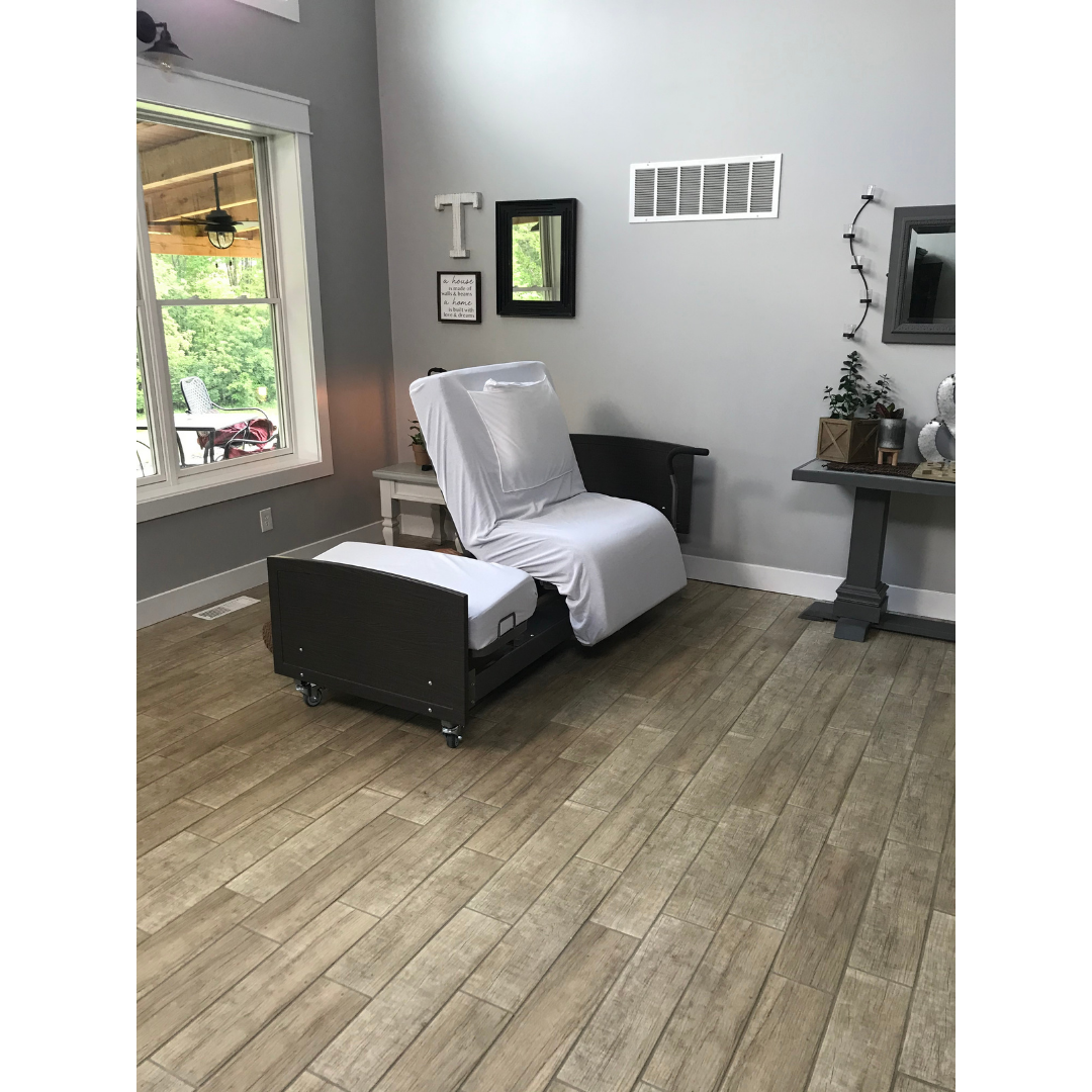 A Med-Mizer ActiveCare Rotating Hospital Bed with white sheets stands in a bright living room featuring wood-style floors, a large window, gray walls adorned with a mirror, artwork, and small plants.