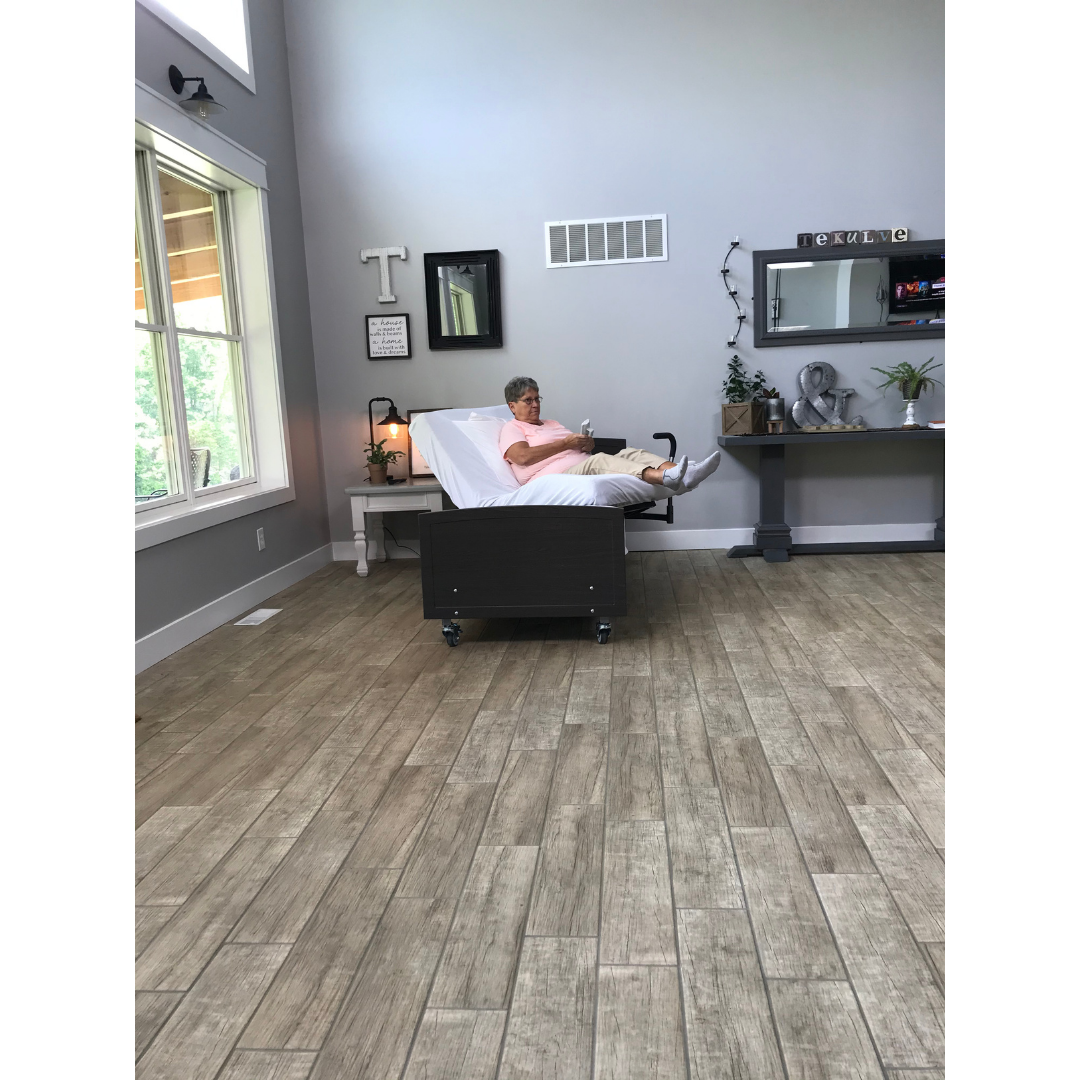 An older woman sits upright in a Med-Mizer ActiveCare Rotating Hospital Bed placed in the center of a spacious, bright room with wood floors, modern decor, large windows, a desk, and wall decorations.