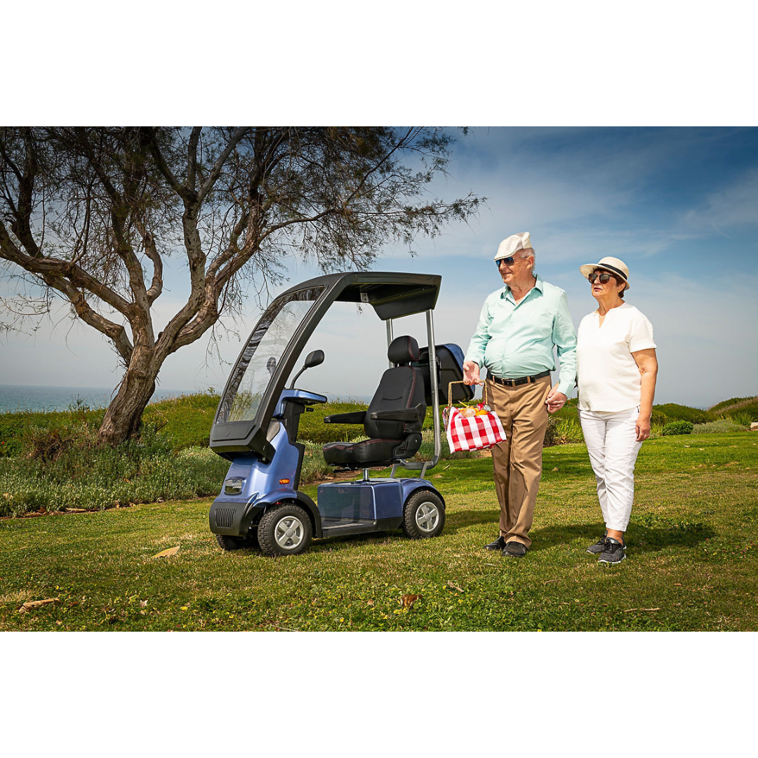 An elderly couple strolls beside the Afikim Afiscooter C4 HD, an electric mobility scooter with a canopy. The man carries a red-checkered bag; both wear hats and sunglasses. An orthopedic seat ensures comfort as they enjoy the scenic view of a tree and blue sky.