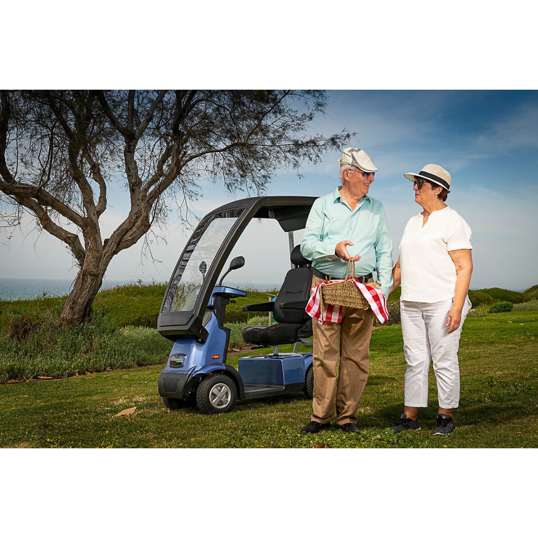 An elderly couple stands on grass near an Afikim Afiscooter C4 HD 4-Wheel Mobility Scooter. The man holds a picnic basket, and both wear sun hats and sunglasses. Behind them, a tree and the sea are visible under a blue sky.