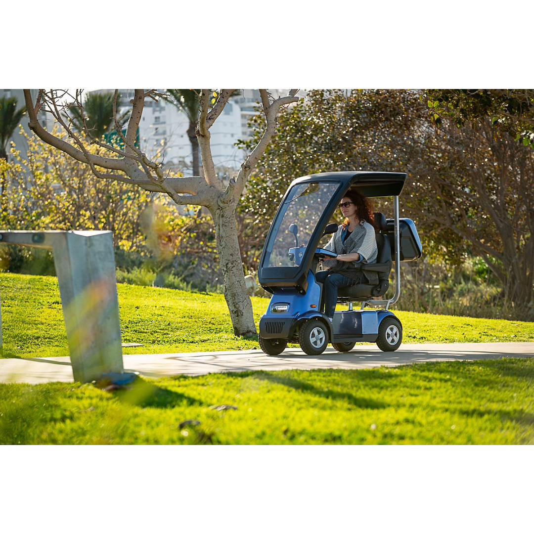 A person rides an Afikim Afiscooter C4 HD 4-Wheel Mobility Scooter with an orthopedic seat along a paved park path, as sunlight filters through trees and casts dappled shadows, while a building looms in the background.