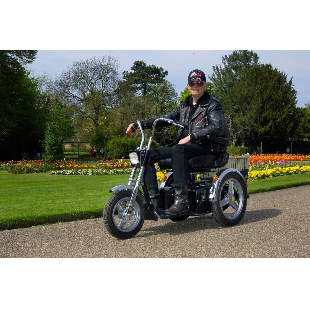 A person in sunglasses and a black leather jacket rides the Afikim Afiscooter SE 3-Wheel Recreational Scooter with bike handles along a garden path. Vibrant flower beds and trees under a clear sky enhance the scenes allure.