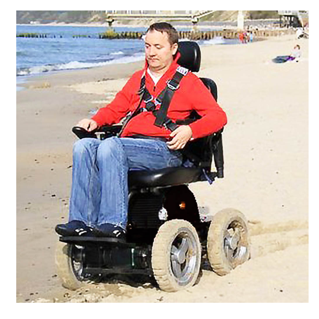 A man in a red shirt and jeans rides a Wheelchair88 All-Terrain Stair Climbing 4x4 Recreational Electric Vehicle with large wheels along a sandy beach near the ocean.