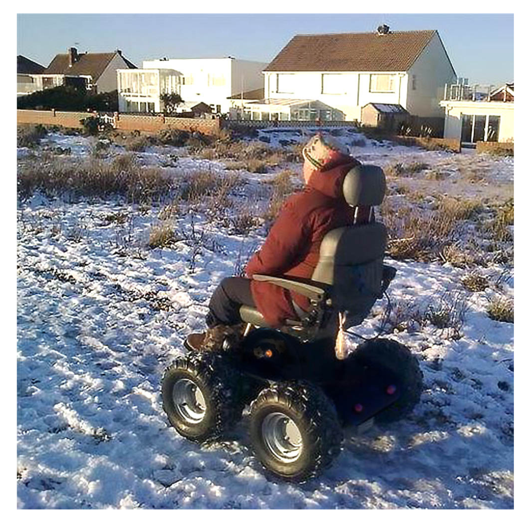 A person in a red coat and hat sits on a Wheelchair88 All-Terrain Stair Climbing 4x4 Recreational Electric Vehicle in a snowy open field with houses visible in the background.
