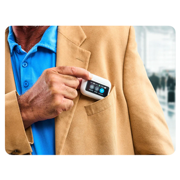 A person in a tan blazer and blue shirt puts an Audien Hearing Atom X Hearing Aid with Digital Case Control, showing app icons on its touchscreen display, into their jacket pocket in an indoor public space.
