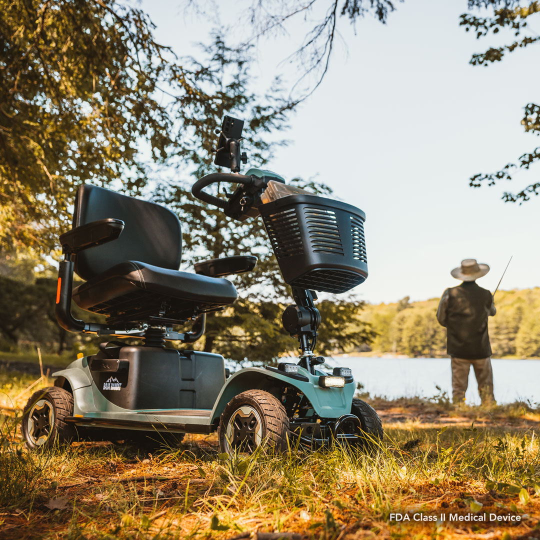 The Pride Baja® Bandit 4 Wheel Scooter, known for its rugged off-road capabilities and full suspension, is parked on grass near a lake. A person in a wide-brimmed hat fishes nearby, surrounded by trees and water, creating a serene outdoor setting.