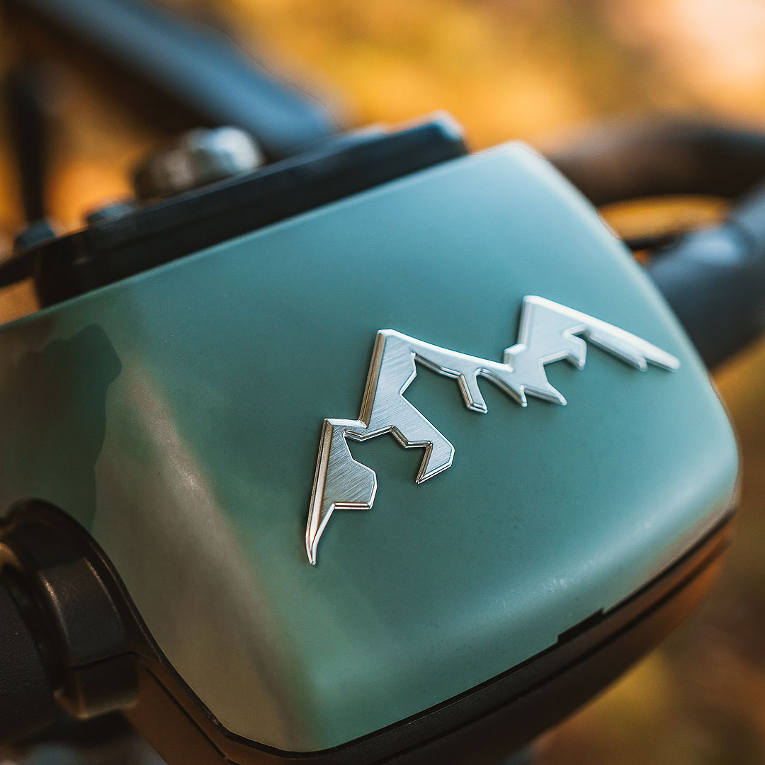 Close-up of a mountain-shaped logo on the green frame of a Pride Baja® Bandit 4 Wheel Scooter, designed for off-road adventures. The metallic, shiny logo contrasts with softly blurred handlebars in warm tones.