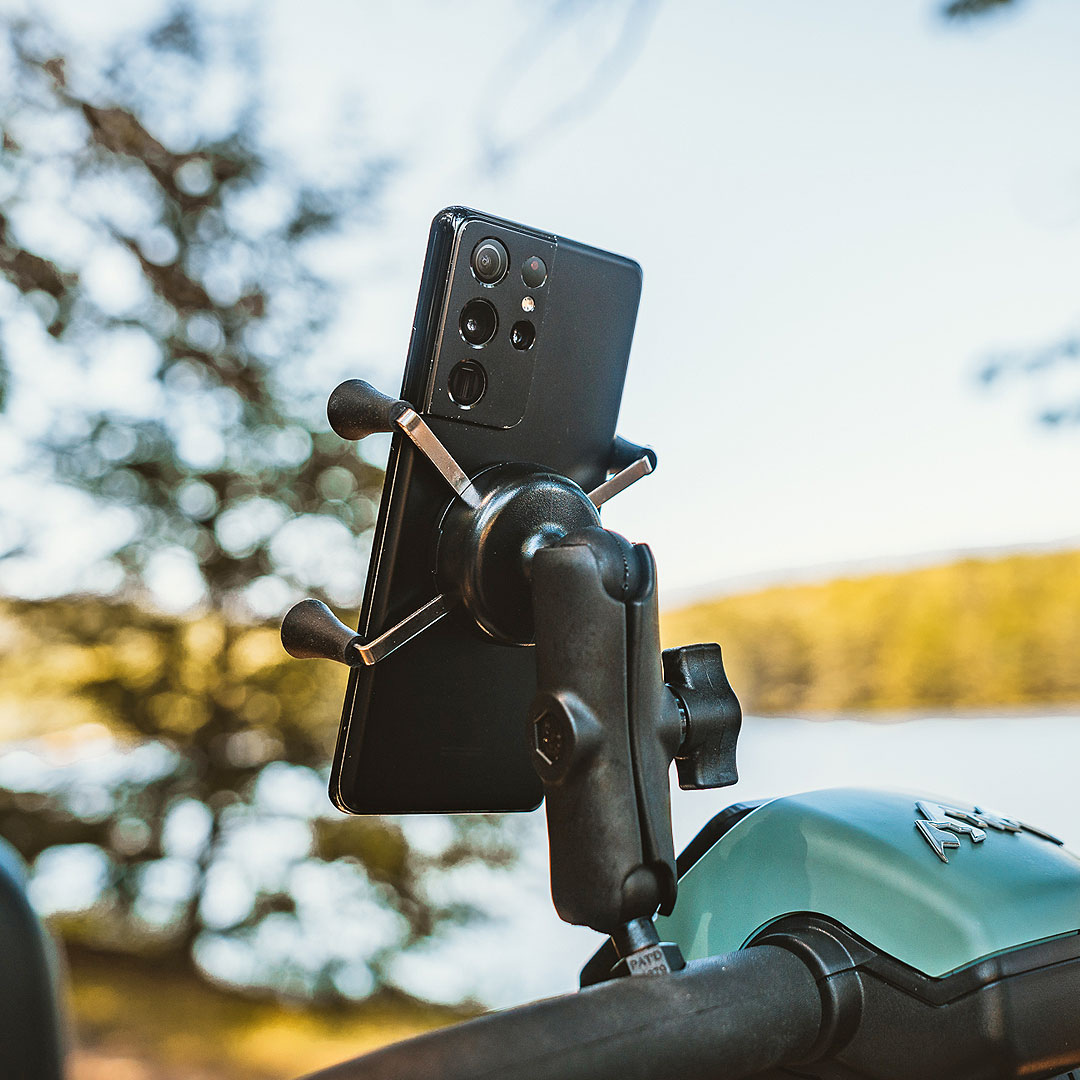 A smartphone mounted on a Pride Baja® Bandit 4 Wheel Scooter handlebar captures the adventurous spirit of an off-road ride. The blurry backdrop of trees and a lake on a sunny day enhances the scene, promising endless exploration.