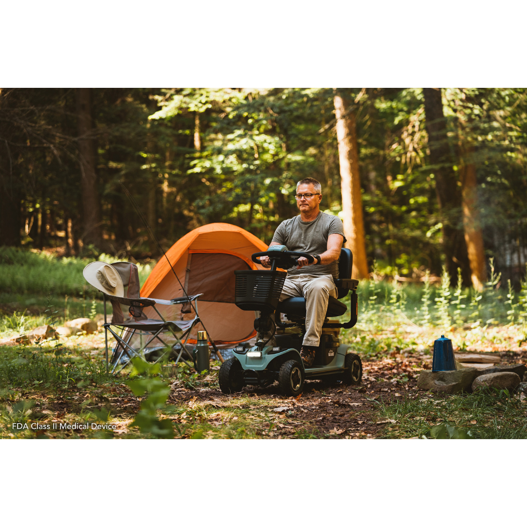 A man sits on a Pride Baja® Bandit 4 Wheel Scooter at a forest campsite. An orange tent is nearby, and a folding chair holds a hat. Sunlight filters through the trees, creating a serene setting perfect for exploring with his full suspension ride.