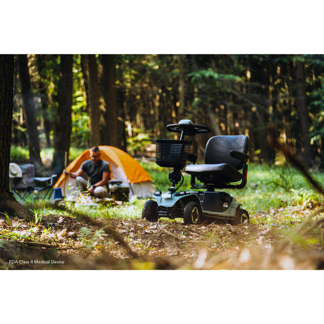A Pride Baja® Bandit 4 Wheel Scooter is parked on a grassy forest area, with a person nearby in front of an orange and white tent. Sunlight filters through the trees, creating a serene outdoor atmosphere enhanced by the scooters full suspension for smooth off-road rides.