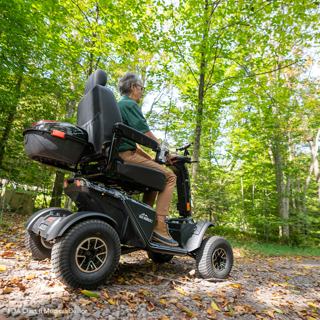 A person navigates a forest trail on the robust Pride Baja® Wrangler® 2, an all-terrain outdoor mobility scooter by Baja Mobility. Surrounded by bright green trees and fallen leaves, it features CTS Suspension for adventurous rides.