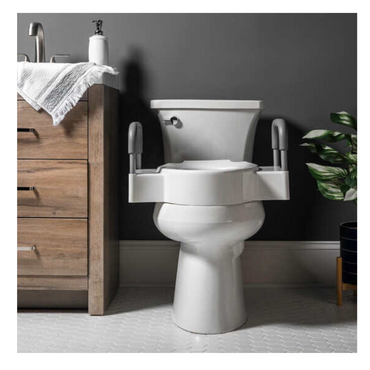 The Bemis Independence Rise™ Elongated/Round 4.5 Raised Toilet Seat with Support Arms is set against a dark gray wall for increased accessibility, next to a wooden cabinet with a silver faucet and white towel. A small plant peeks in on the right, and the floors light geometric pattern adds warmth.