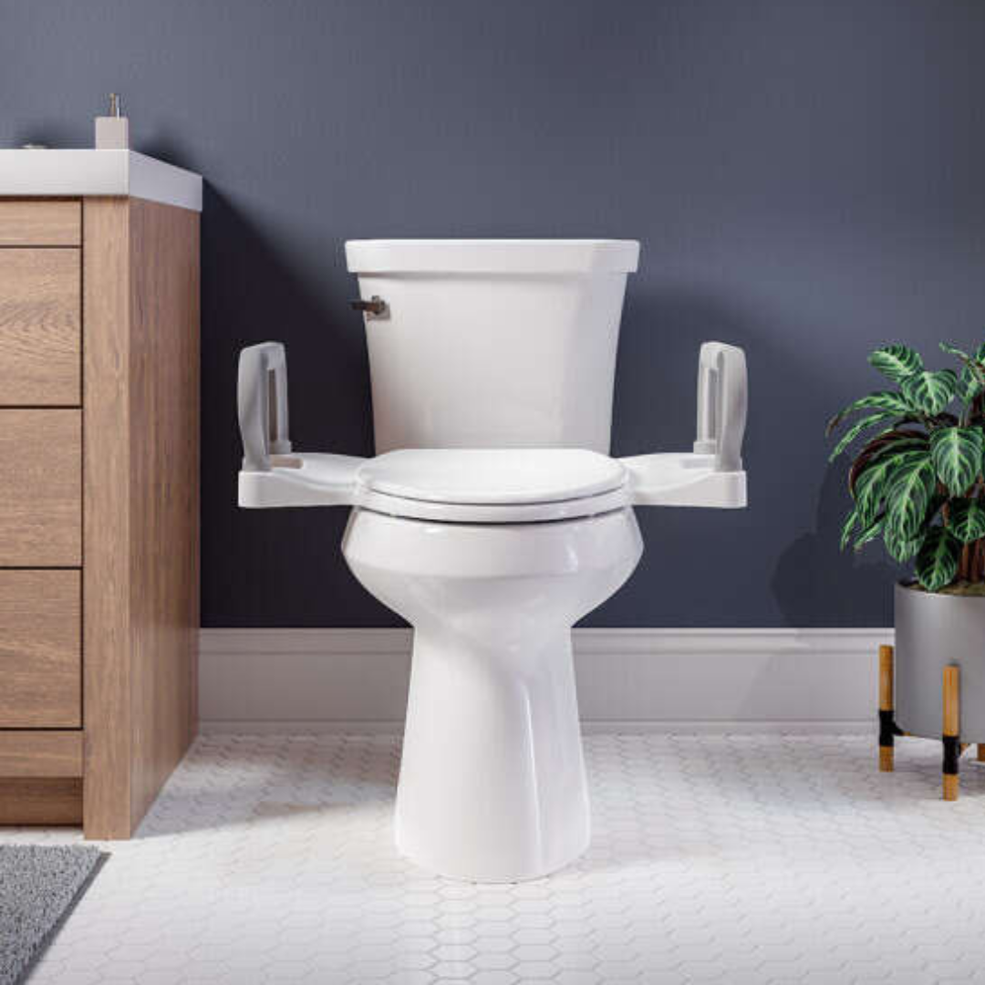 A white toilet featuring the Bemis Assist™ Standard Height Premium Toilet Seat with supportive arms sits in the bathroom. To the left is a wooden cabinet, while a green potted plant adorns the right, all against a dark gray wall and sleek white hexagonal tile floor.
