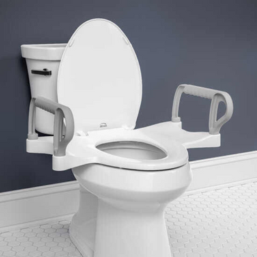 A bathroom features a Bemis Assist™ Standard Height Premium Toilet Seat with raised design and support arms, set against dark gray walls and light hexagonal tile flooring. The Stay·Tite Installation System ensures secure setup, while the lid remains open.