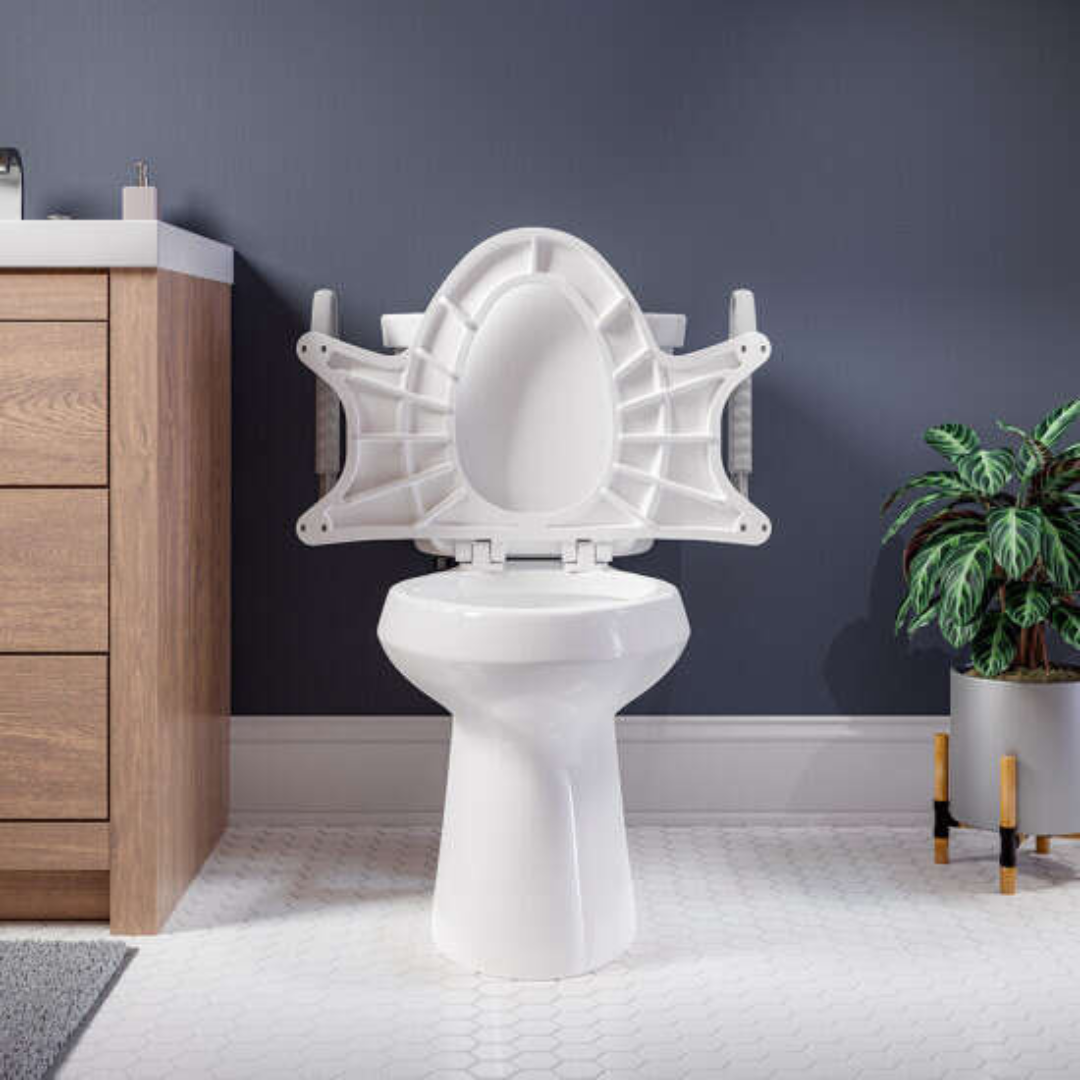 A Bemis Assist™ white toilet with its standard height seat raised highlights the underside, set against a dark gray wall. On the left is a wood-finished vanity, and to the right, a potted plant. The floor features hexagonal white tiles, completing this Stay·Tite Installation System equipped bathroom scene.