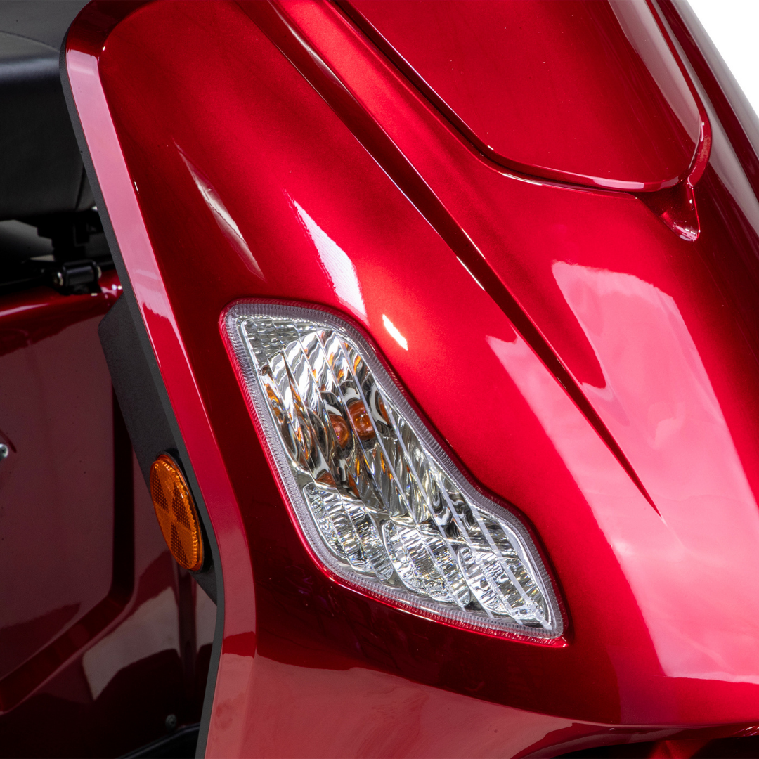 Close-up of the Bespoke Legacy Recreational 3-Wheel Scooter’s shiny red front, with a clear headlight and orange side reflector from its electric lighting system, reflecting light against a white background.
