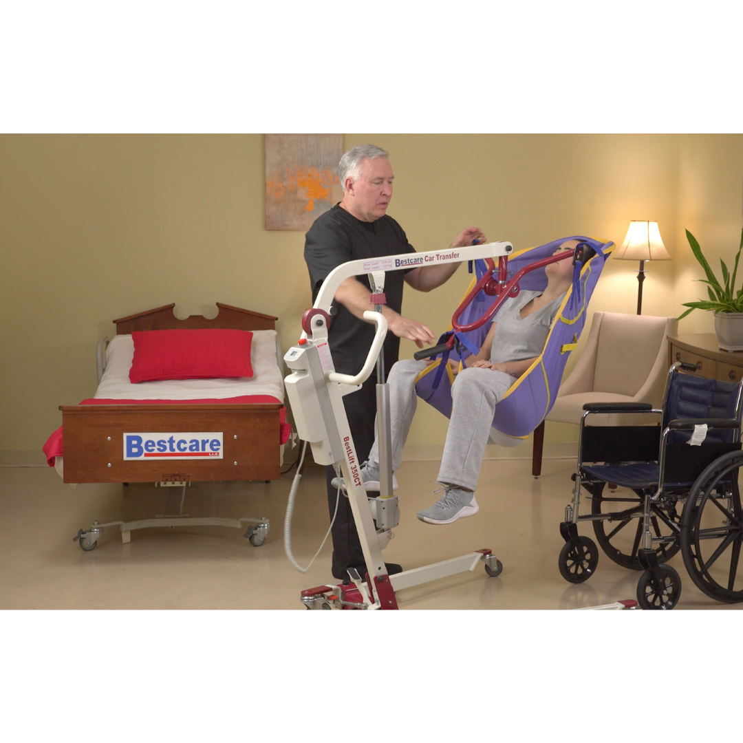 A man uses the Bestcare BestLift PL350CT Folding Portable Patient Lift to transfer an elderly person from a wheelchair to a bed. The room includes a red-accented bed, a plant, and a chair, all catering perfectly to mobility needs.