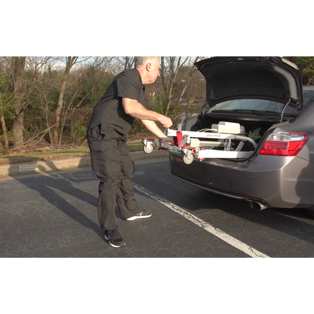 A person in black clothing lifts a Bestcare BestLift PL350CT Folding Portable Patient Lift from a grey cars trunk in a parking lot, enhancing mobility. Trees in the background add nature to this moment of car transfers and accessibility.