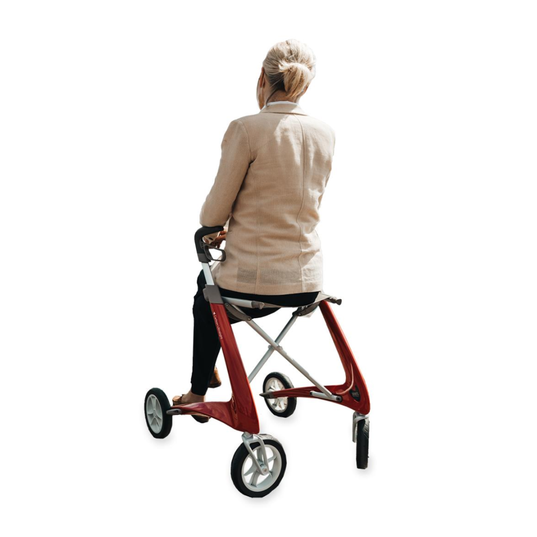 Seen from behind, a blonde woman sits on the ByACRE Ultralight Carbon Fiber Rollator - 10.6 lbs, with its minimalist red frame and ergonomic handles. Her beige jacket and black pants complement the rollators design featuring large wheels, all set against a plain white background.