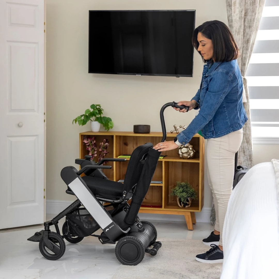 A woman in a denim jacket and light pants stands indoors beside a bed, holding the handle of a WHILL Model F powerchair equipped with a Cane Holder; a TV and wooden shelf are visible in the background.