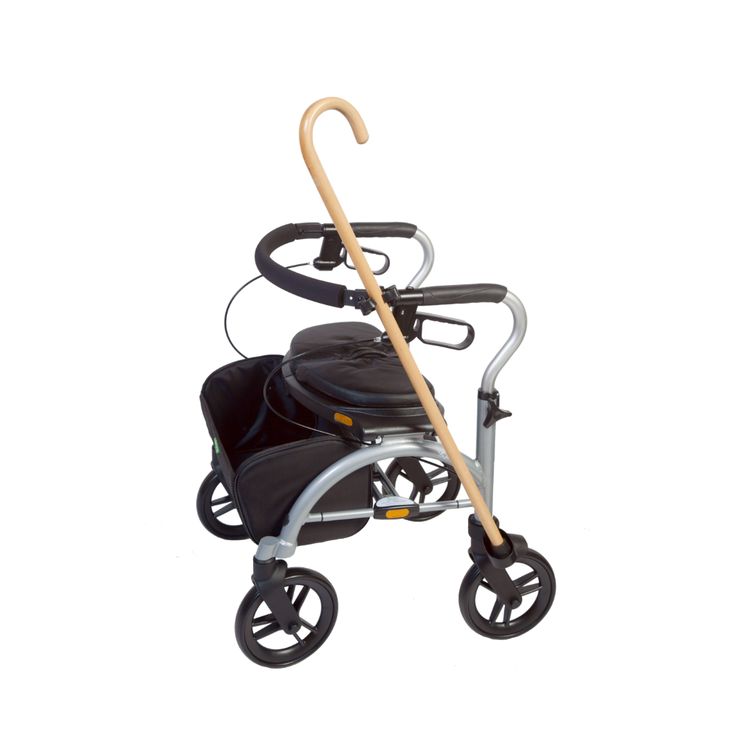 The Cane Holder for Evolution Rollators securely attaches to a silver walker with four wheels, black seat, and storage basket, allowing a light wood cane to rest diagonally for easy access.