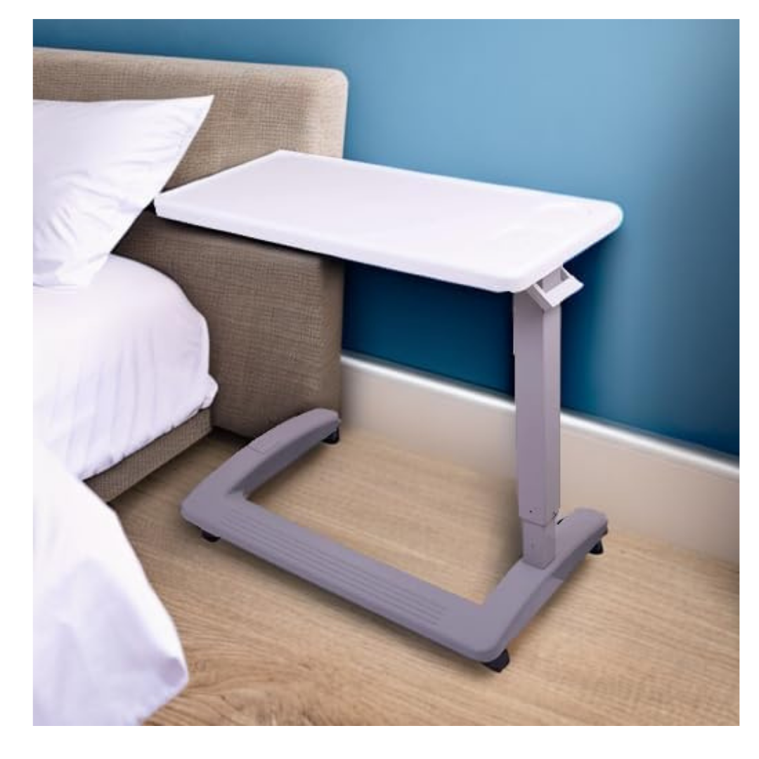 The Carex Height Adjustable Rolling Overbed Table with XL Surface, featuring a white top and gray base on wheels, is placed beside a bed with a white pillow against a blue wall on a wooden floor.