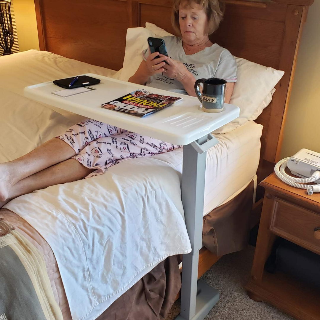 An older woman sits up in bed using her phone, with a Carex Height Adjustable Rolling Overbed Table with XL Surface in front of her holding a magazine, coffee mug, and computer mouse. Medical equipment is on the bedside table.