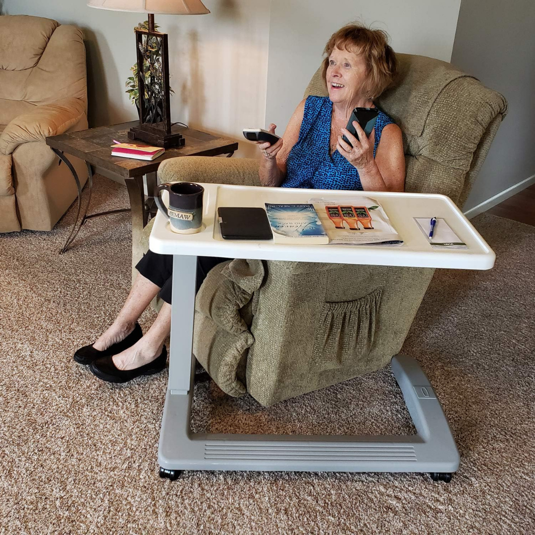 An older woman smiles in an armchair, holding a remote and phone. In front of her is a Carex Height Adjustable Rolling Overbed Table with XL Surface, topped with coffee, a tablet, magazines, and a pen. A side table and lamp are nearby on the carpeted floor.