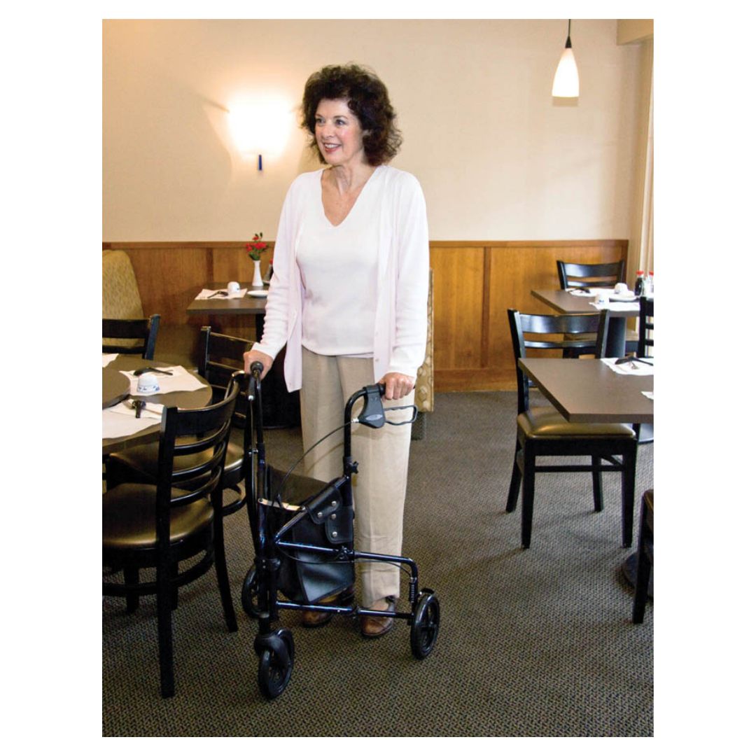 A woman with curly hair stands smiling in a restaurant, holding her Carex Trio Roller 3-Wheel Walker Rollator. She wears a white top, light pink cardigan, and beige pants. Tables and chairs are arranged around her.
