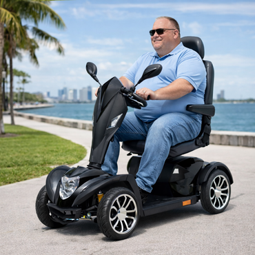 A man in sunglasses and a light blue polo shirt rides the Drive Cobra GT4 Heavy Duty Bariatric Mobility Scooter along a grassy waterfront path, with palm trees and a city skyline in the background.