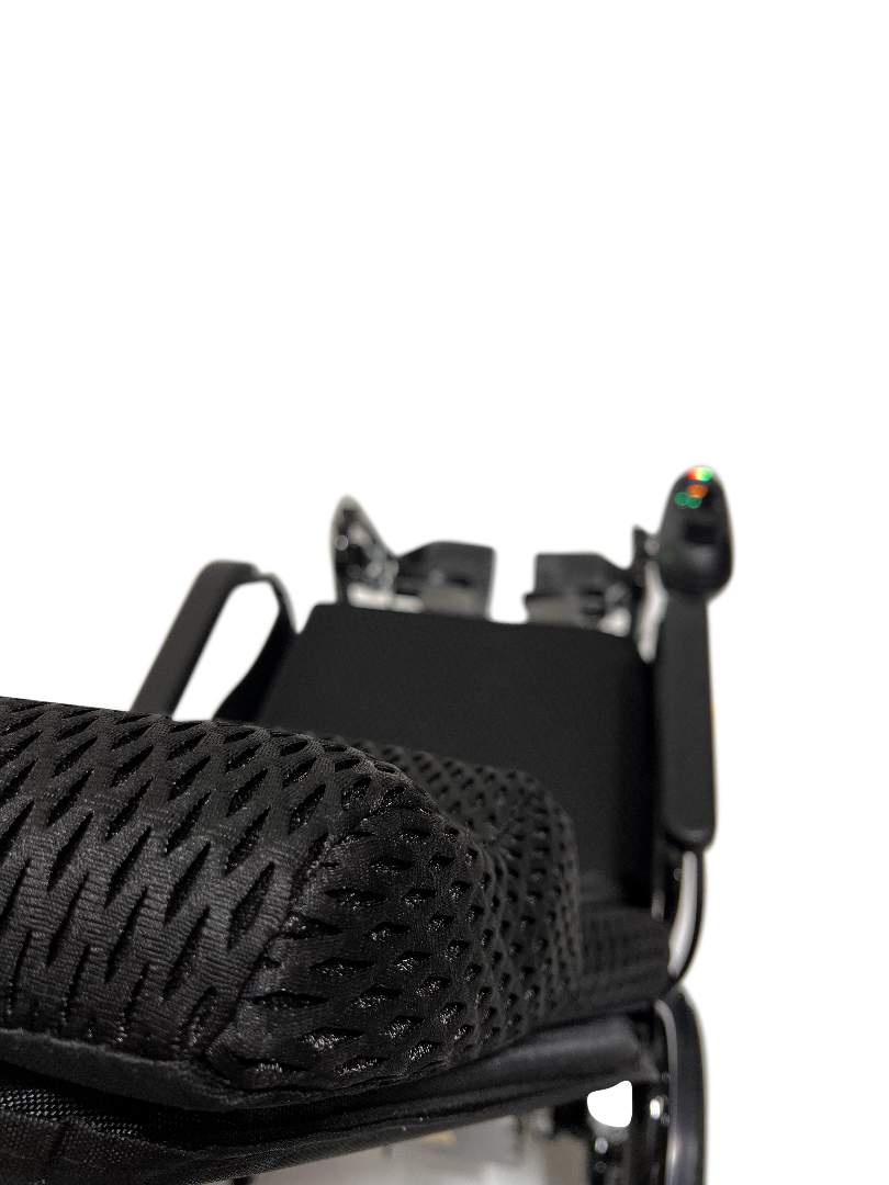 Close-up of the black patterned seat cushion on the ComfyGo X-9 Remote Controlled Pwerchair - Automatic Reclining, showcasing perforations for breathability. The armrest and control panel with a joystick are partially visible on the right.