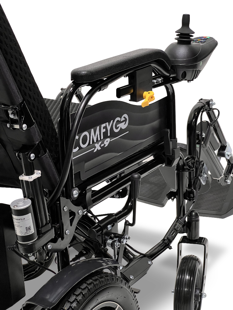 Close-up of a black ComfyGo X-9 Remote Controlled Powerchair - Automatic Reclining, featuring the joystick on the armrest. Its detailed mechanical frame and rear wheel highlight precision engineering in this innovative mobility solution.