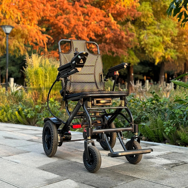 The EASWE B10 Portable Folding Powerchair - Airline Approved, a modern black electric wheelchair with dual motors, mesh seating, and sturdy wheels, sits empty on a park path surrounded by autumn trees and lush greenery.